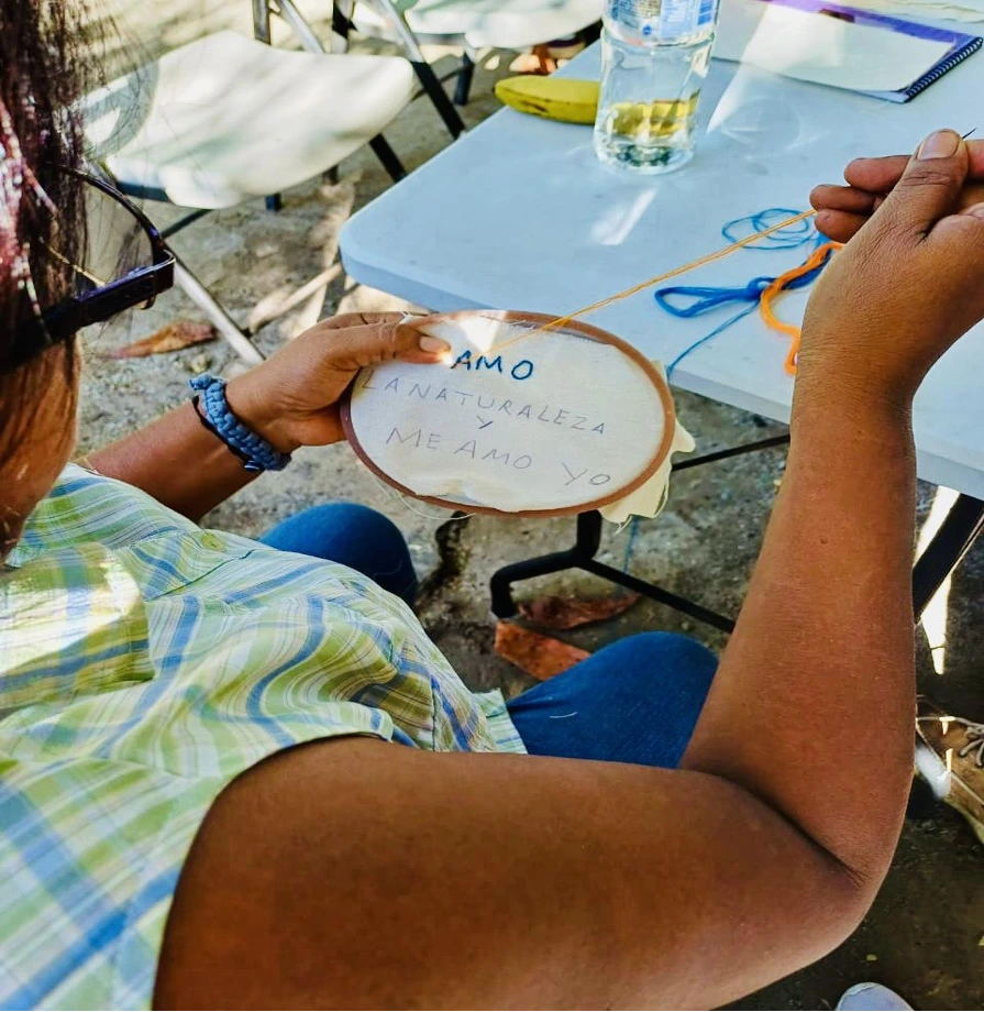 Person embroidering the phrase “I LOVE NATURE AND I LOVE MYSELF” on a fabric hoop, sitting outdoors with colorful threads, a bottle of water, and a banana on the table.