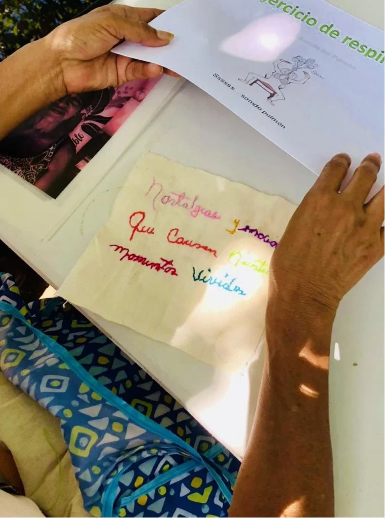 Person holding a sheet titled “Breathing Exercise” featuring an illustration and the text “SSSSS lung sound”; on the table, there is an embroidery with the phrase “Nostalgias and emotions caused by lived moments” and a black-and-white photo of a person wearing a hat.